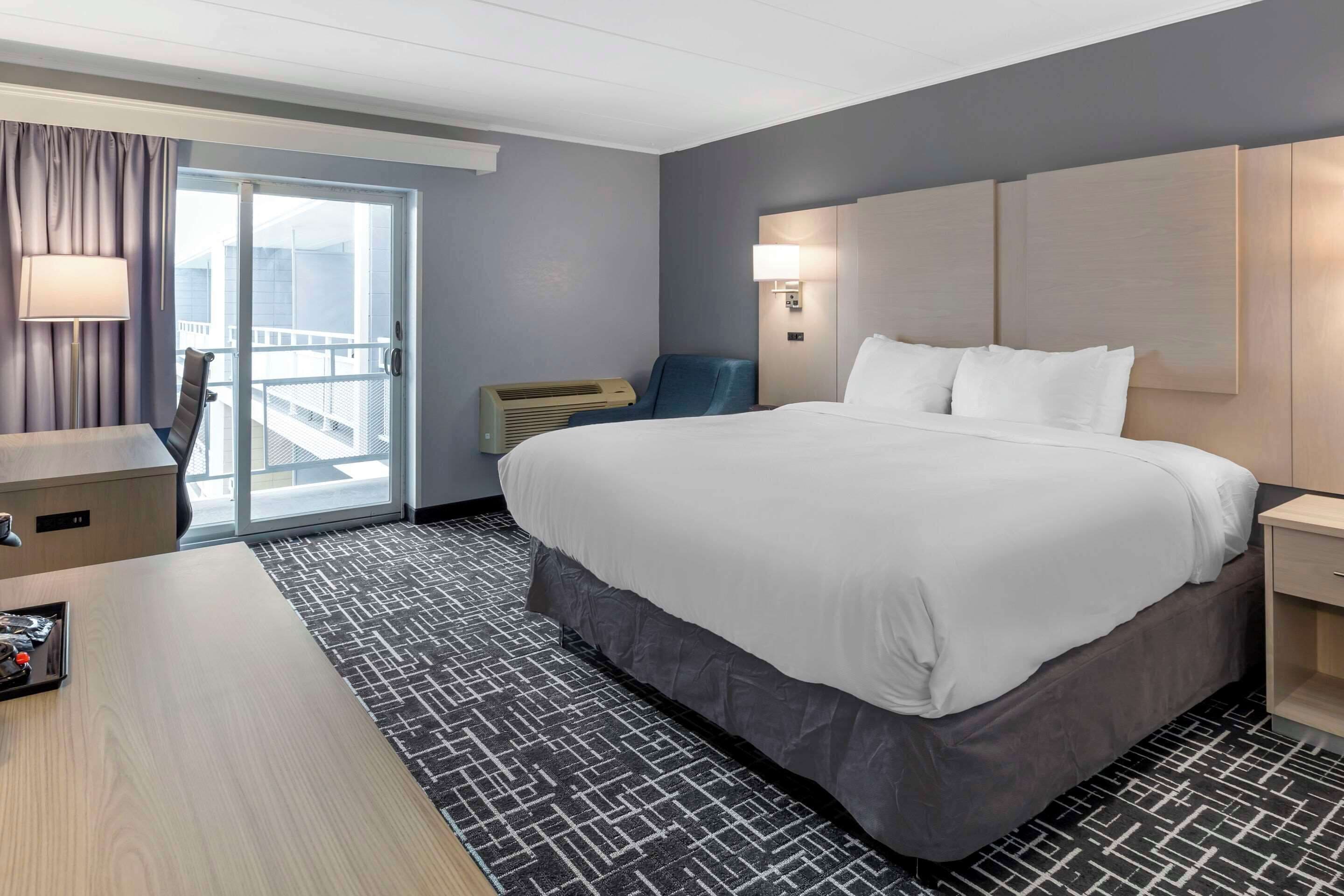 A hotel room with a large bed, a desk, a chair, a lamp, a wall-mounted air conditioner, and a sliding glass door to the balcony.