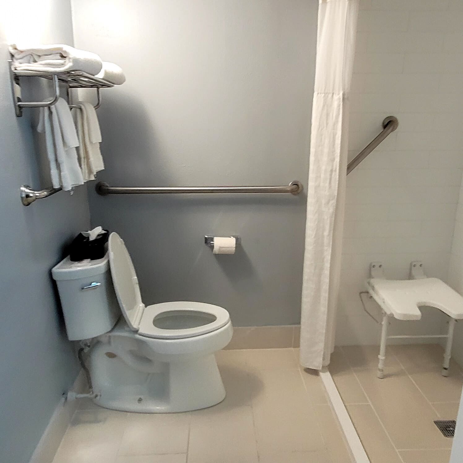 Un baño con inodoro, barra de apoyo, toallero y un pequeño banco.