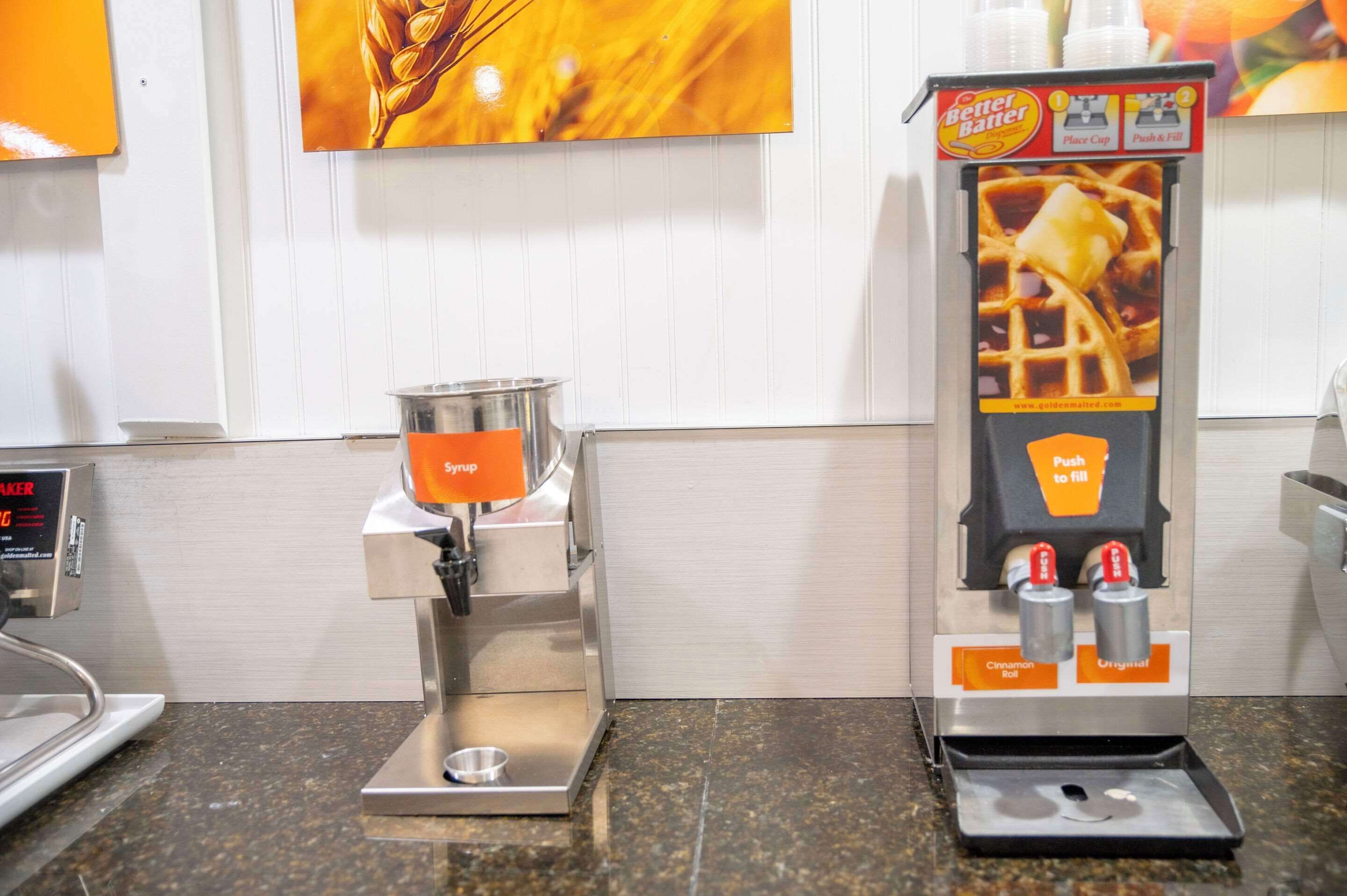 A syrup dispenser with a push-to-fill mechanism, a coffee maker, and a wall-mounted advertisement.