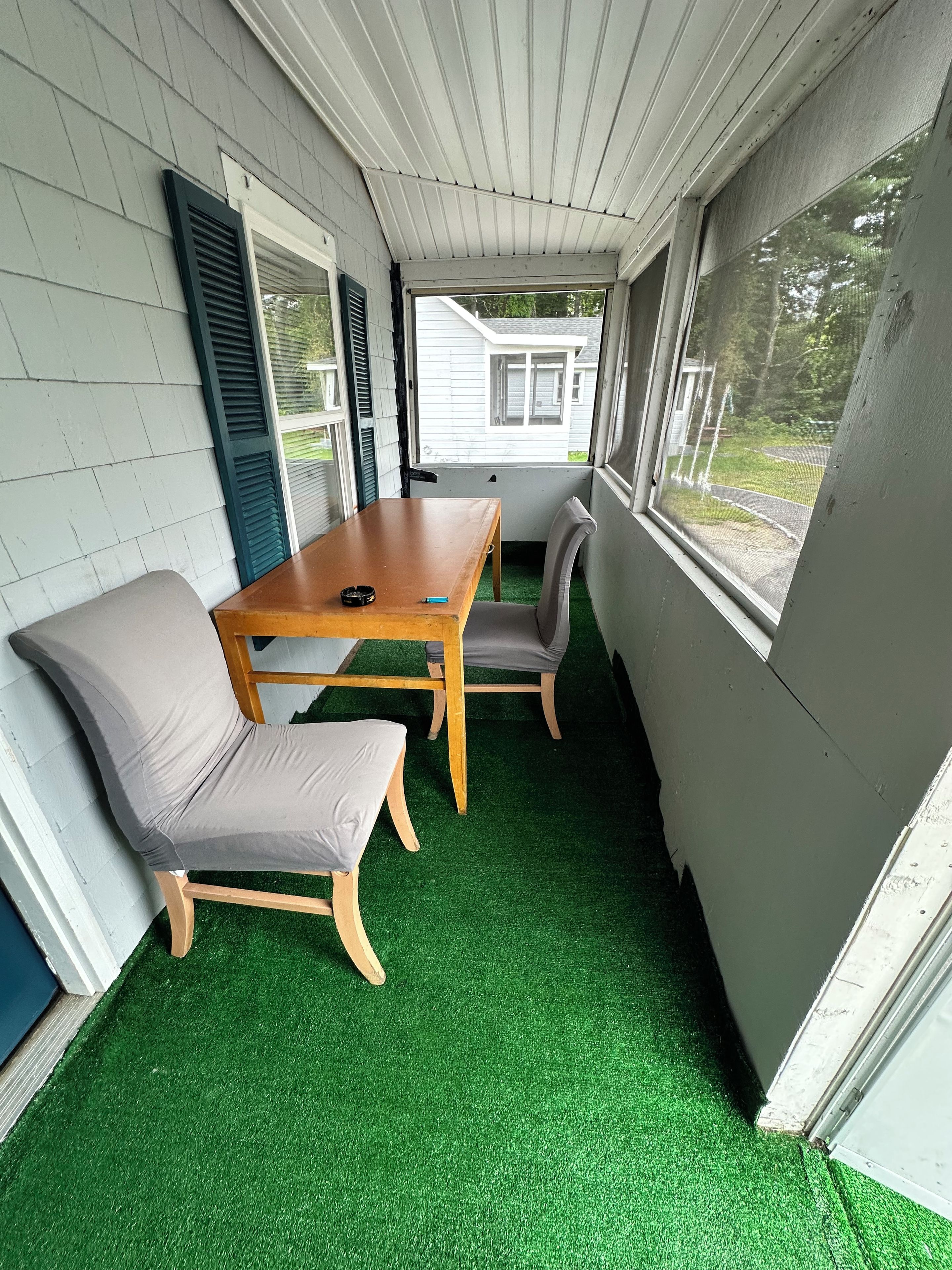 Een overdekt terras met een houten tafel en twee stoelen, een raam met uitzicht op een huis, en een vloer van kunstgras.