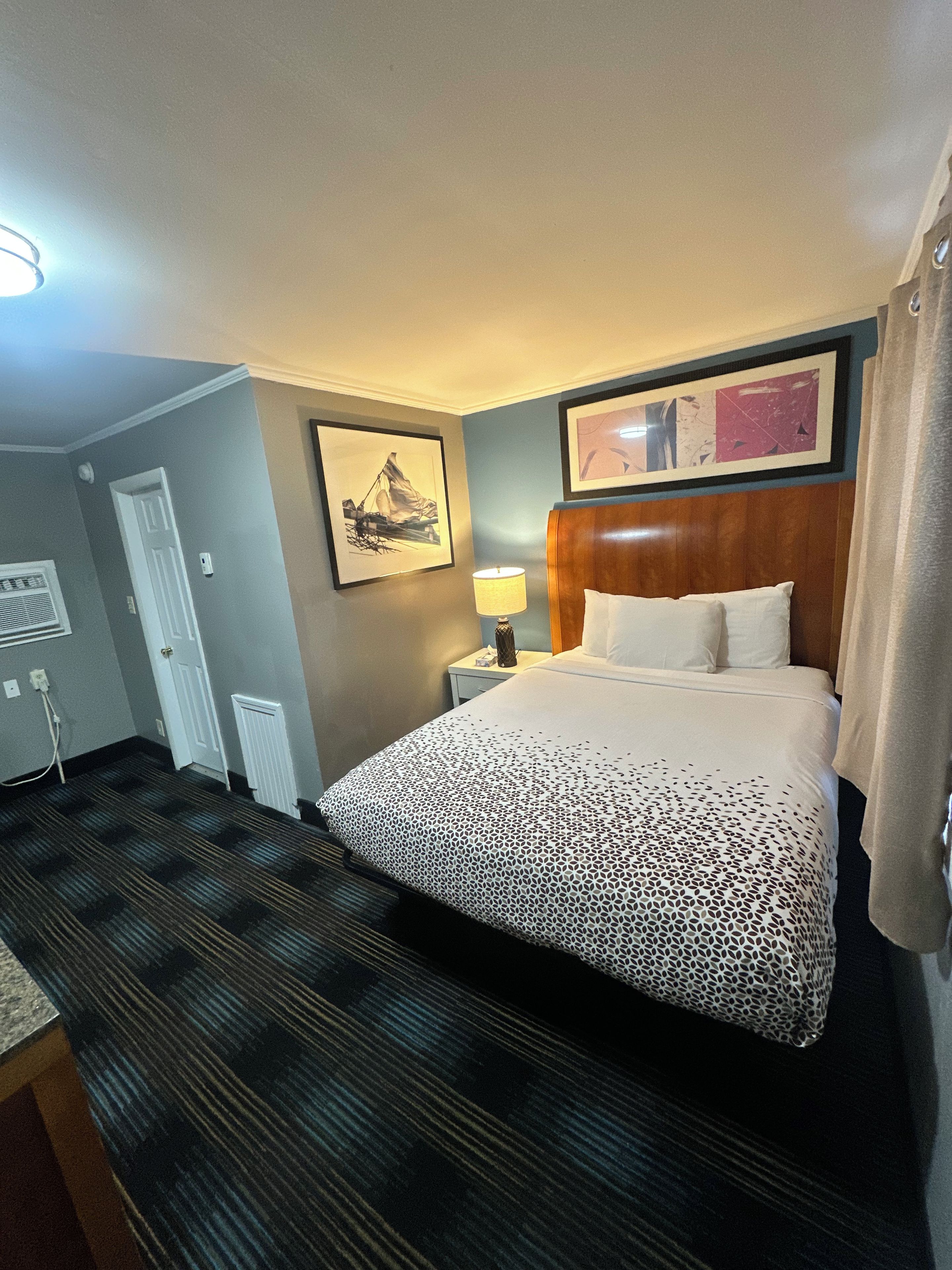 A hotel room with a bed, a nightstand, a lamp, a framed picture on the wall, and a door.