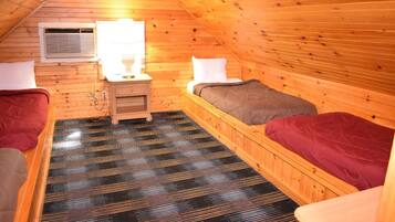 A cozy attic bedroom with wooden walls and a sloped ceiling. It includes two beds, a nightstand, and a lamp.