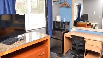 A compact hotel room with a kitchenette, a wooden desk, a chair, a television, a microwave, and a refrigerator.