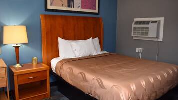 A hotel room with a wooden headboard, a bed with a brown patterned bedspread, a nightstand with a lamp, and an air conditioning unit on the wall.