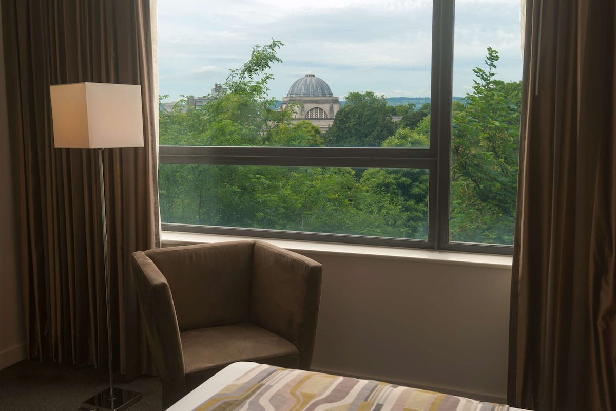 Quarto de hotel com uma janela grande, uma cadeira e um abajur.