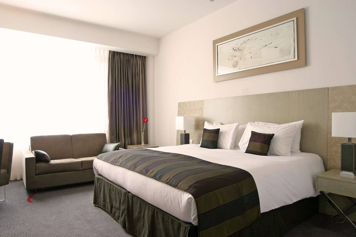 Quarto de hotel com uma cama grande, um sofá, uma mesa de cabeceira com abajur e um quadro na parede.