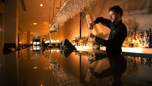Un espace bar doté d’un comptoir aux surfaces polies et réfléchissantes, un barman préparant un cocktail et, en arrière-plan, une variété de bouteilles.