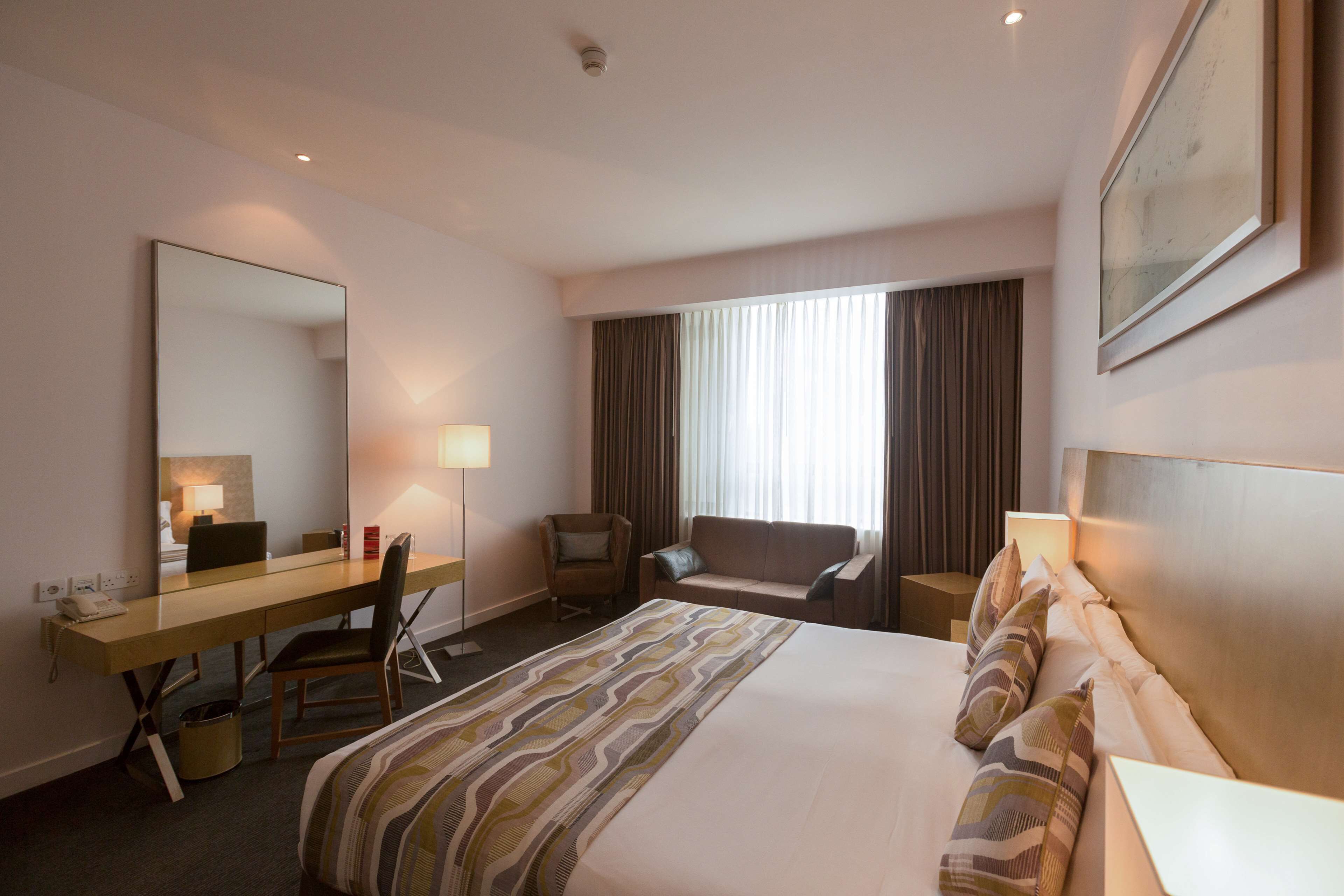 A hotel room with a large bed, a desk, a chair, a sofa, and a large mirror.