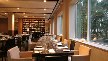 En restaurang med ett långt bord dukat för en måltid, ett vinkällare i bakgrunden och en utsikt över träd genom fönstret.