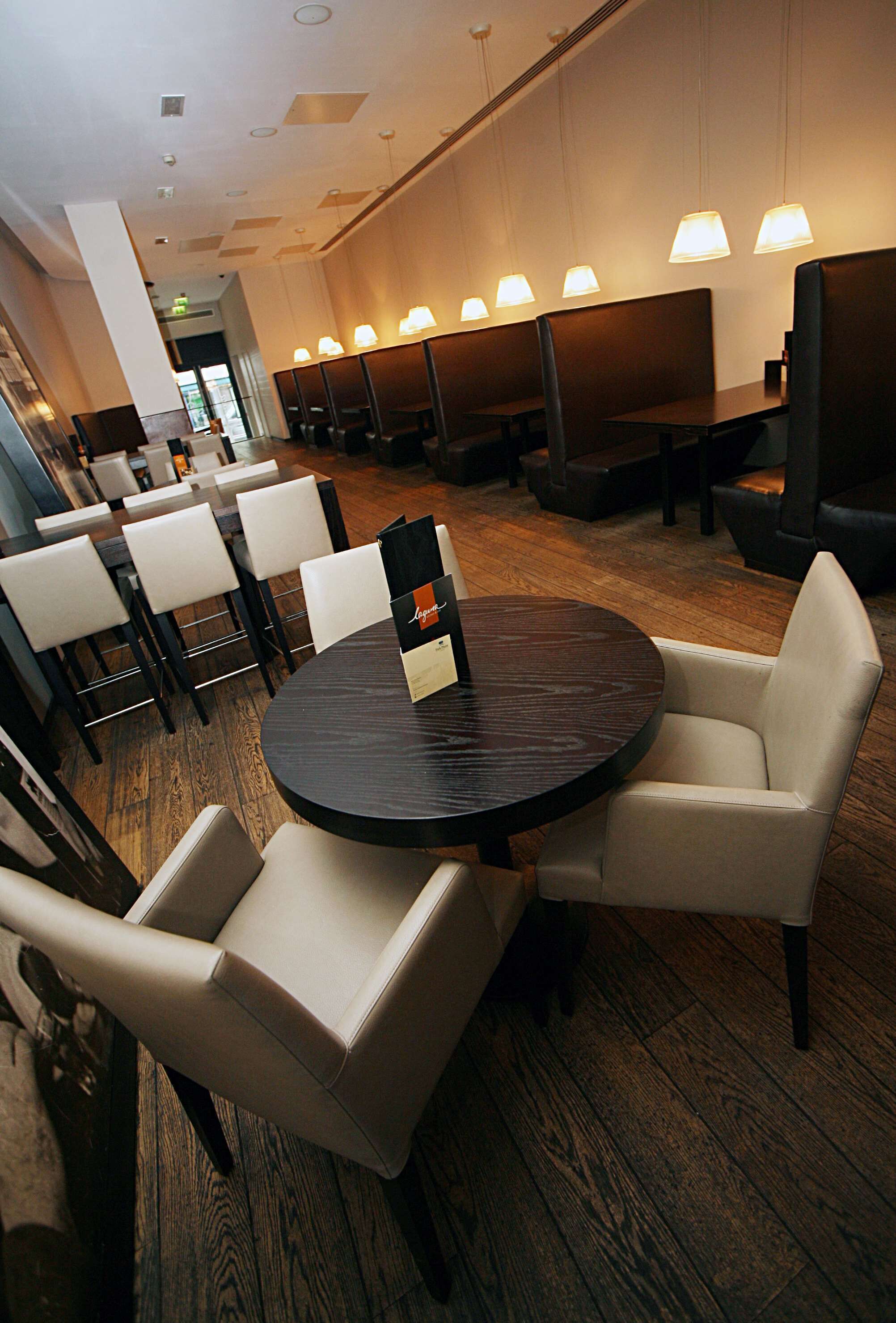 A modern restaurant with round tables and cushioned chairs, wooden flooring, and pendant lighting.