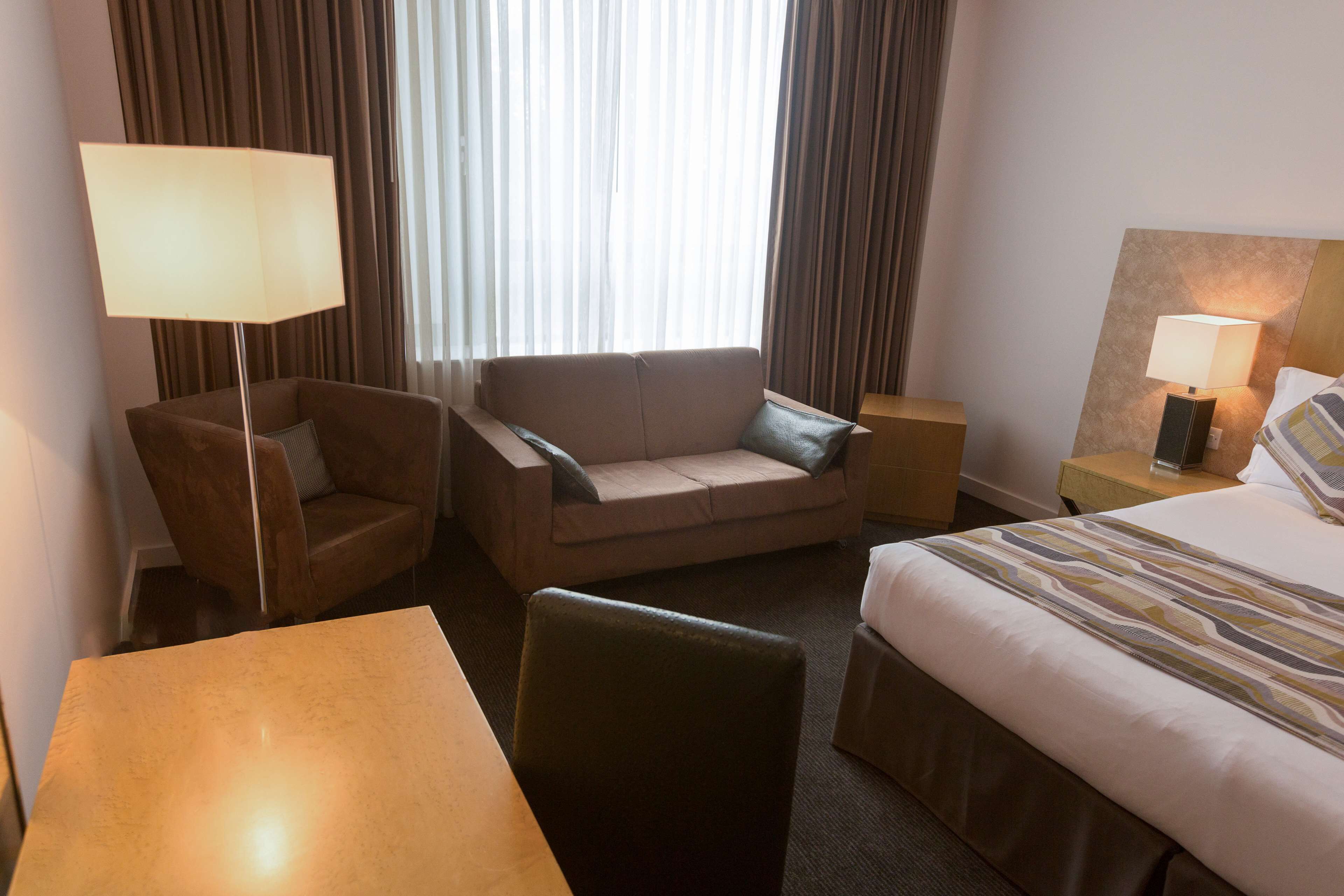 A hotel room with a bed, a sofa, a chair, a table, and a lamp.