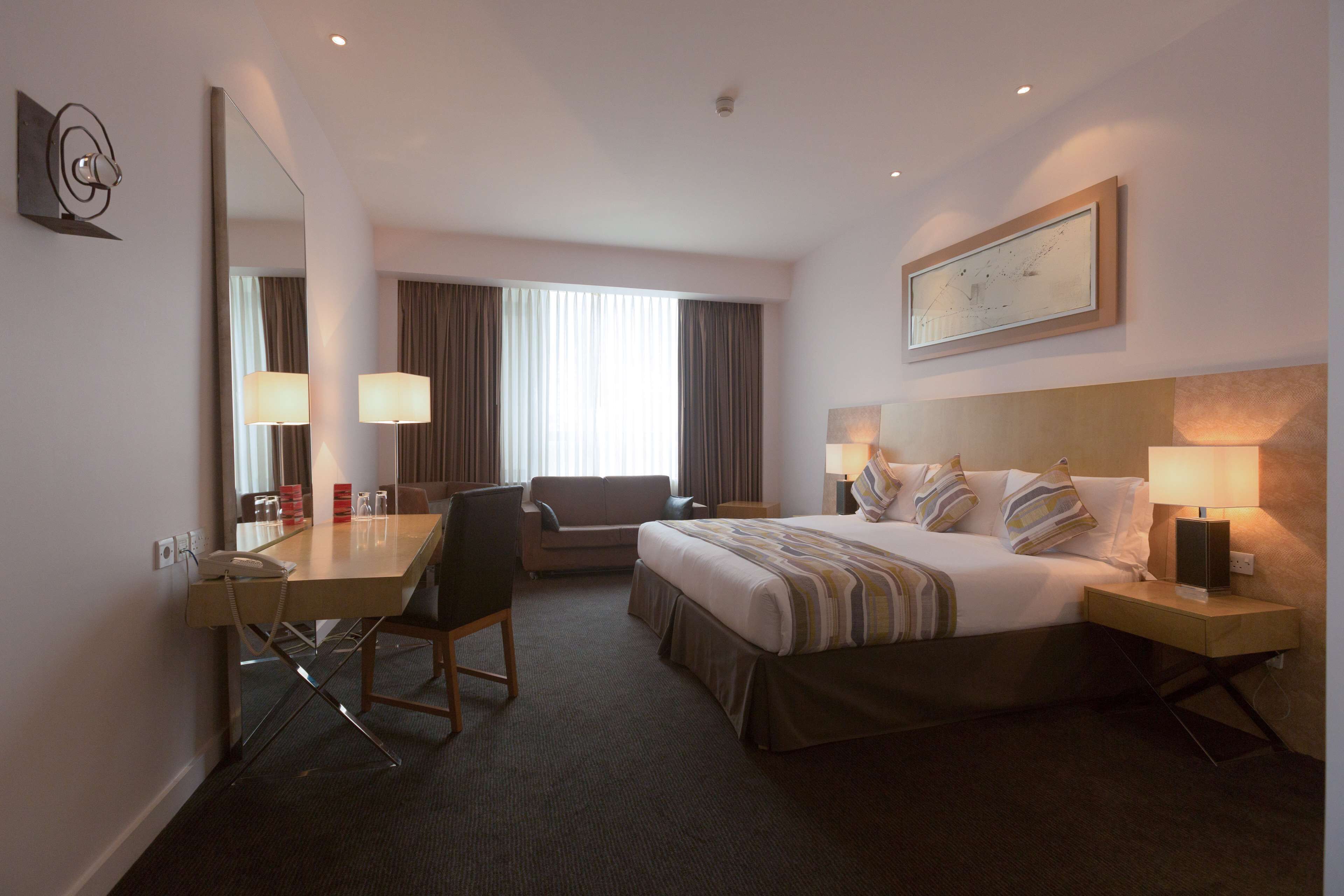 A hotel room with a bed, a desk, a chair, a bedside table, a lamp, a mirror, and a window with curtains.