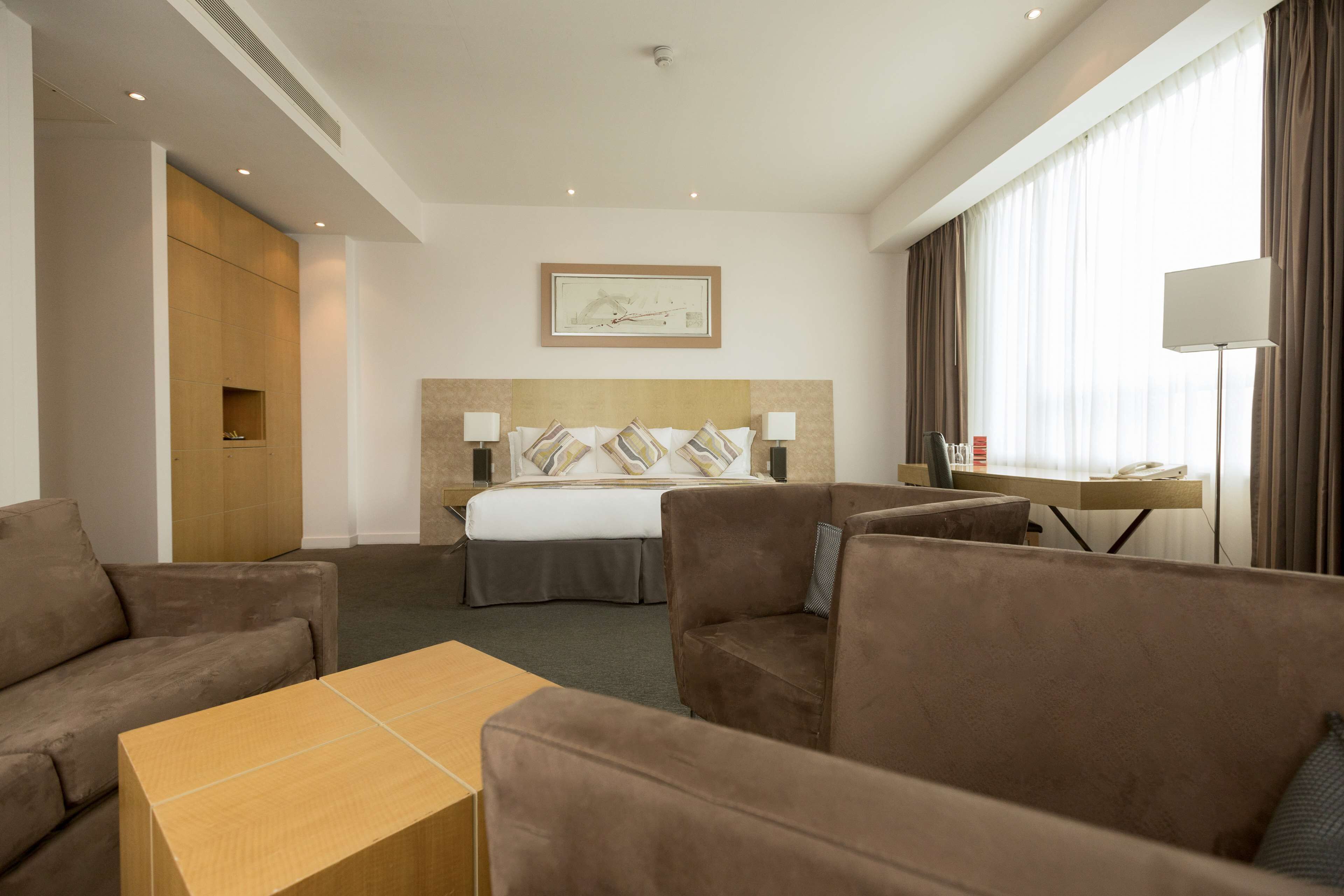 Una habitación de hotel con una cama, dos sillones, una mesita, un escritorio con lámpara y un cuadro en la pared.