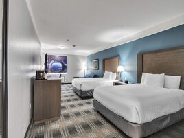 Quarto de hotel com duas camas, uma escrivaninha, uma cafeteira, uma janela com vista para o espaço e uma parede azul.