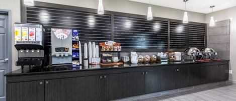 A modern kitchen area with a coffee machine, tea dispenser, and a variety of food items on the counter.