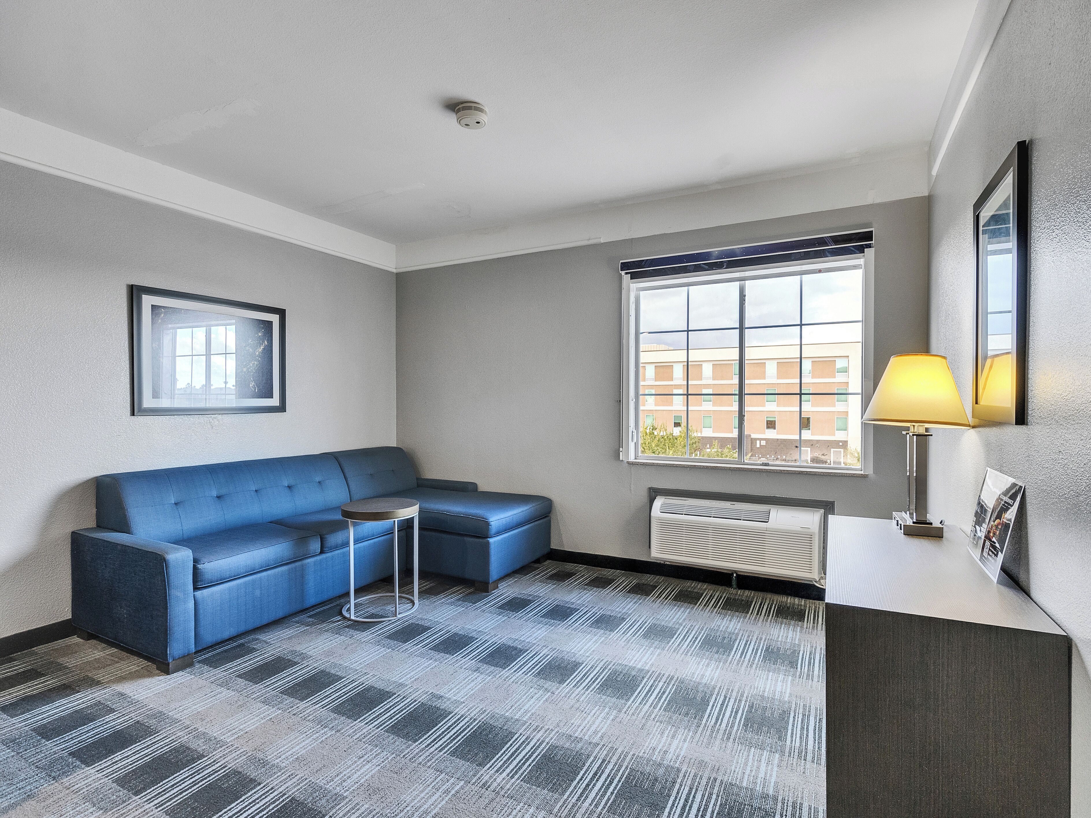 Una habitación de hotel moderna con un sofá azul, una pequeña mesa redonda, un ventanal con vistas a edificios y un radiador.