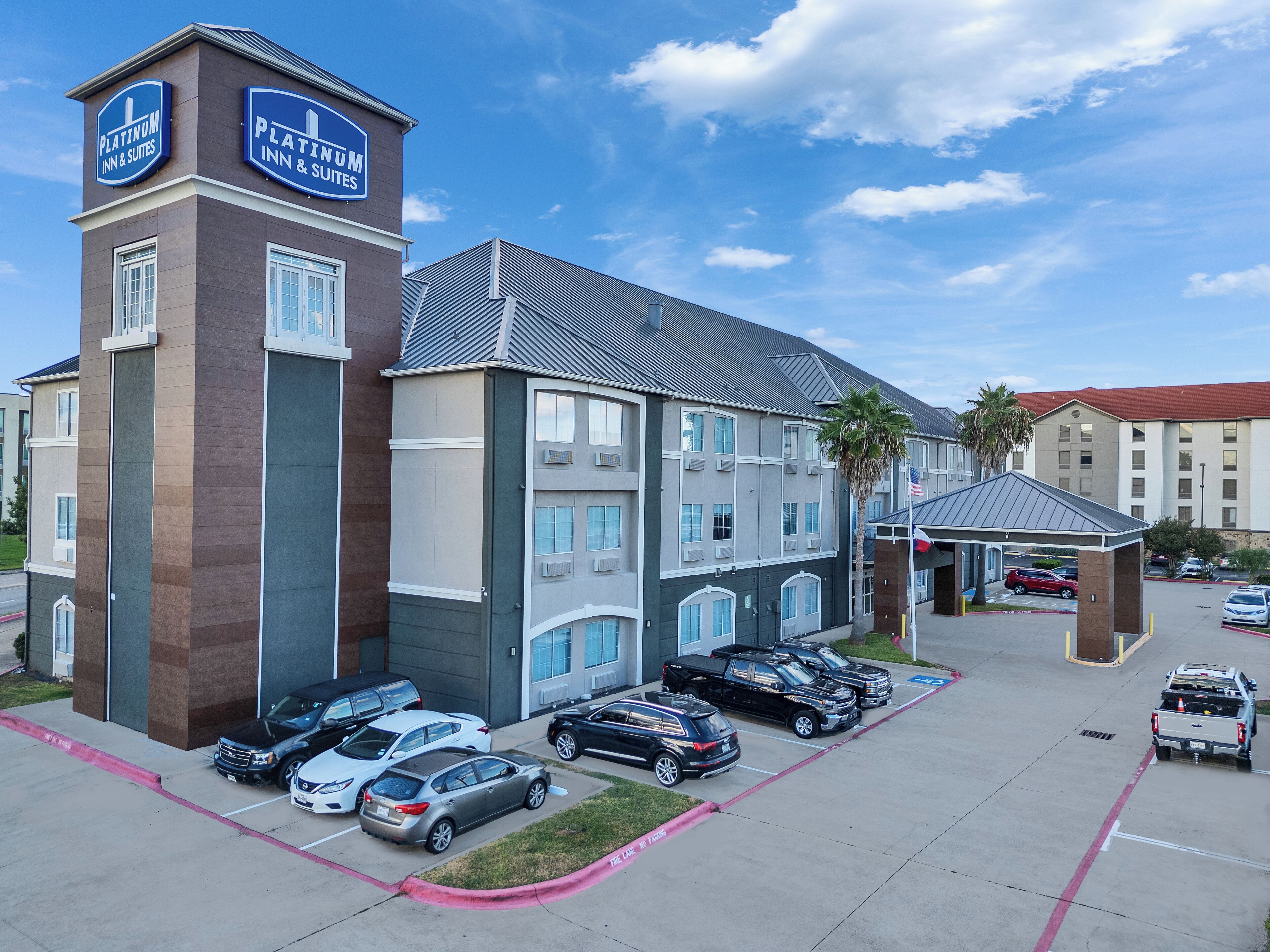 Un hotel con un llamativo letrero de Platinum Inn & Suites, que presenta un diseño arquitectónico moderno con una combinación de fachada de ladrillo y color gris, varios espacios de estacionamiento y una pequeña zona cubierta con la bandera estadounidense.