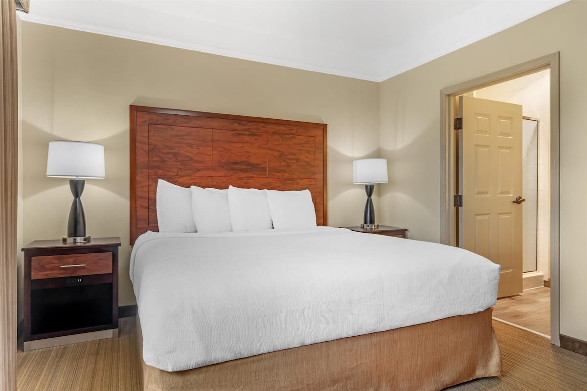 Une chambre d’hôtel avec un grand lit, deux tables de chevet avec des lampes, une tête de lit en bois et une porte donnant sur une autre pièce.