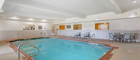 Una piscina interior con superficie de agua cristalina, una escalera de piscina y un borde alicatado. Hay sillas de descanso y una mesa con sillas alrededor de la zona de la piscina.