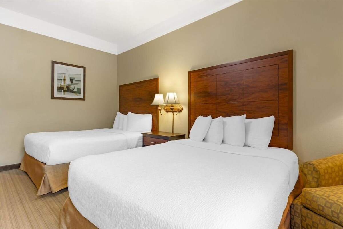 A hotel room with two beds, a nightstand with a lamp, a framed picture on the wall, and a patterned armchair.