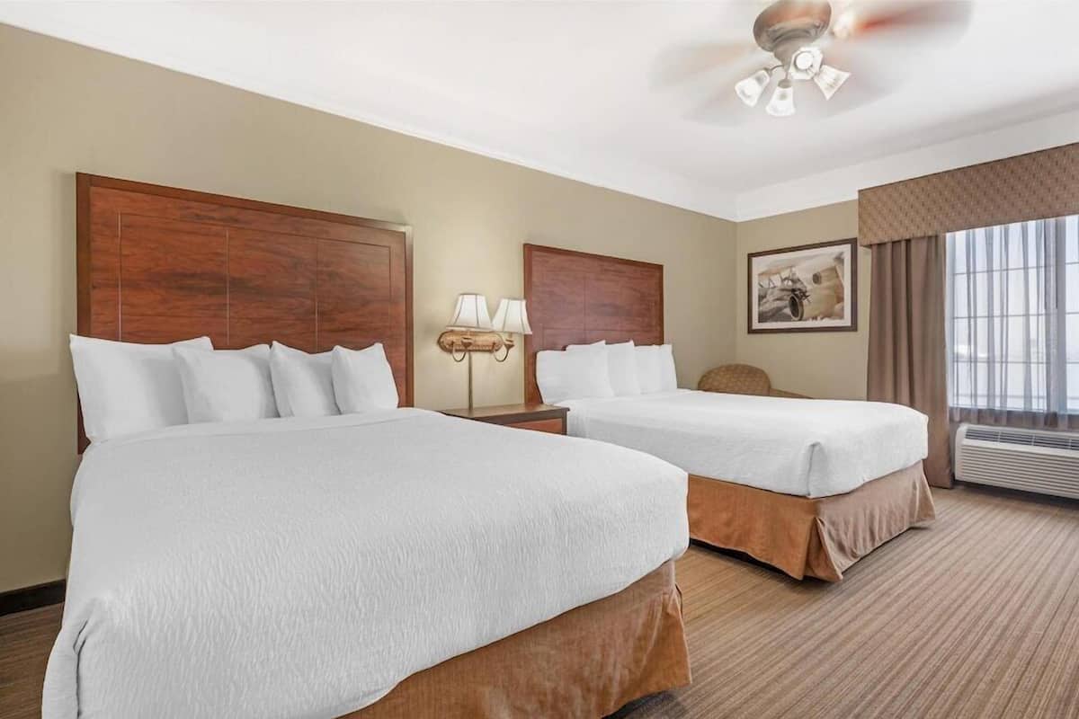 Une chambre d’hôtel avec deux lits, un ventilateur de plafond, une lampe fixée au mur et une grande fenêtre avec des rideaux.