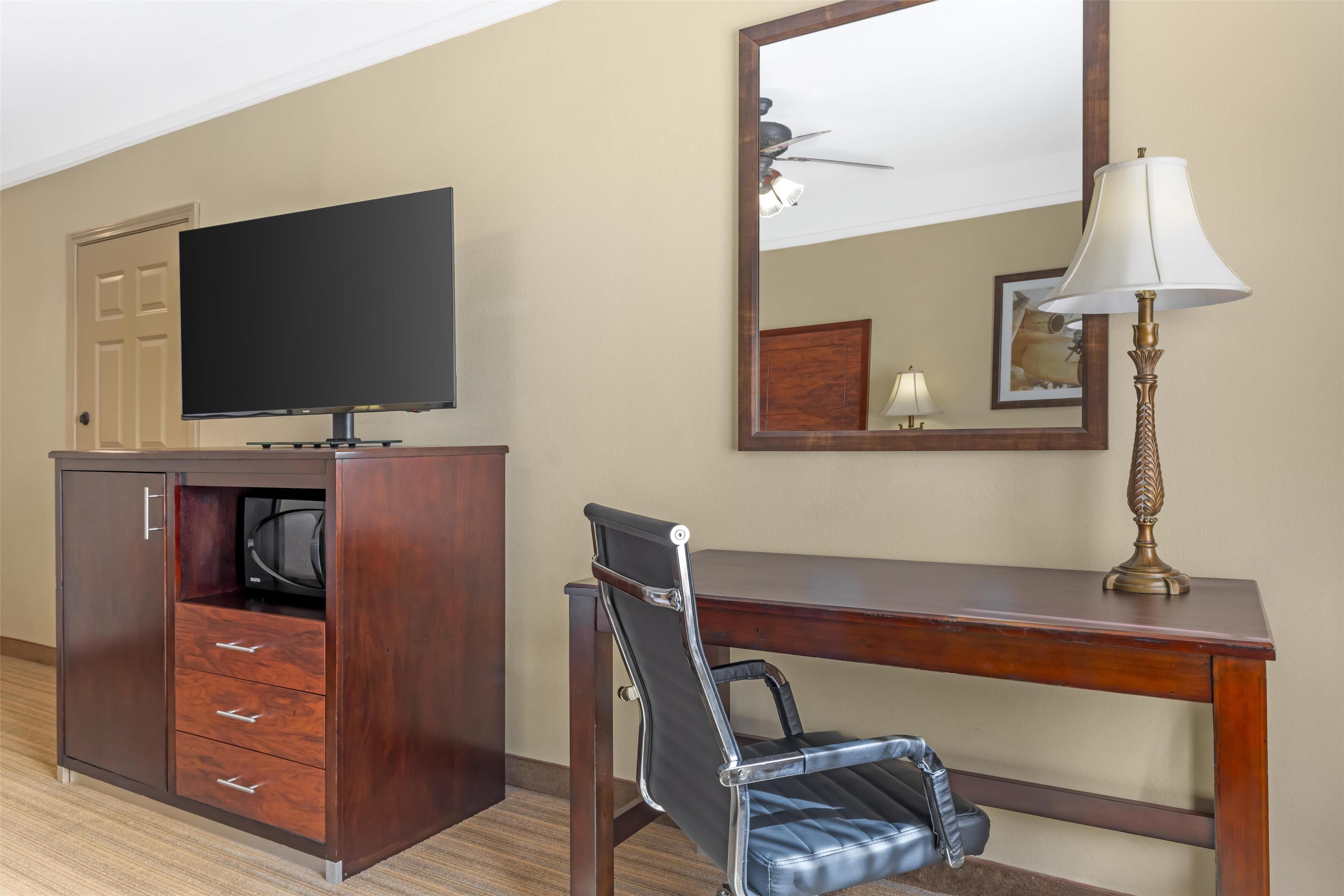 Habitación de hotel con escritorio de madera, una silla, televisor, espejo y lámpara.