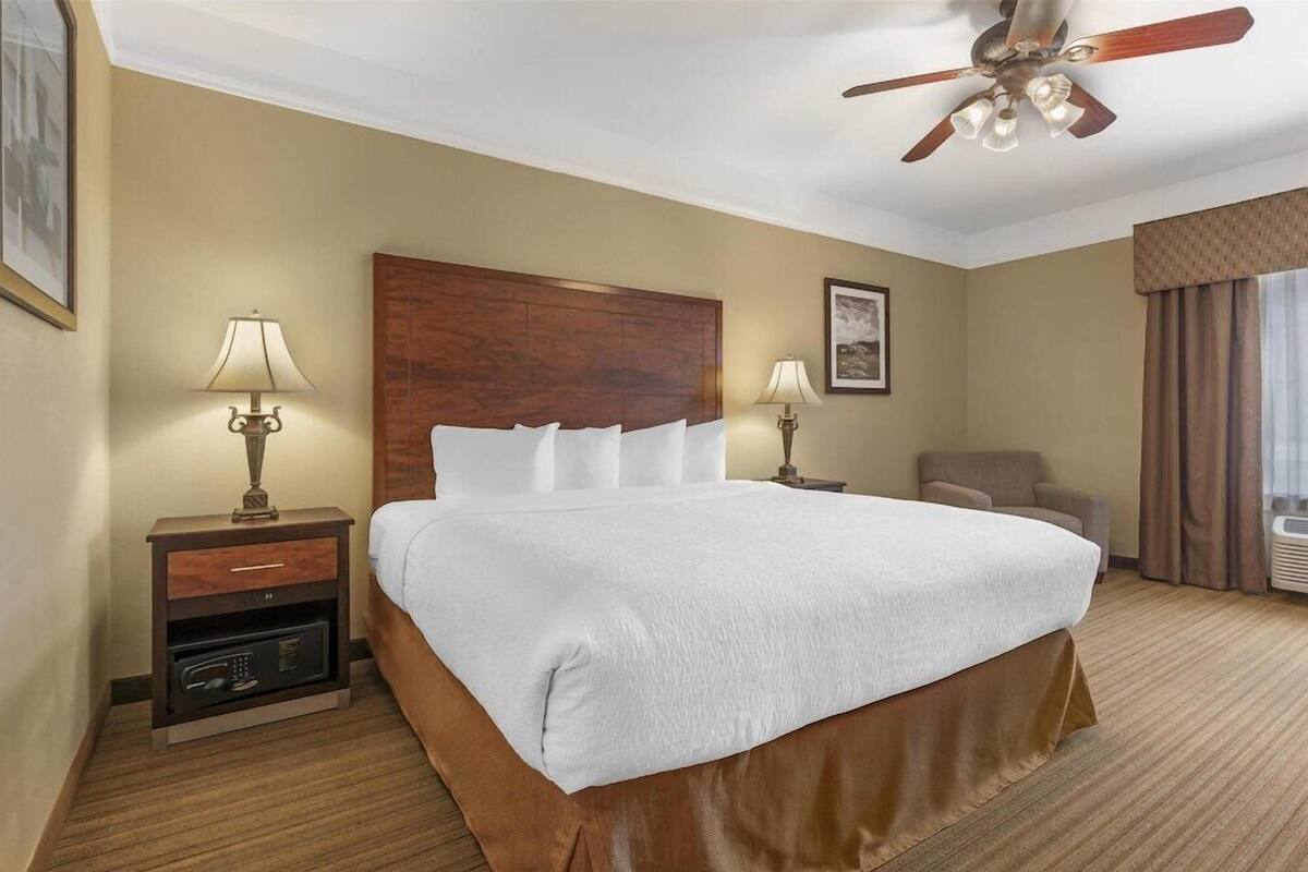 Une chambre d’hôtel avec un grand lit, deux tables de chevet avec des lampes, un ventilateur de plafond, une chaise et une fenêtre avec des rideaux.