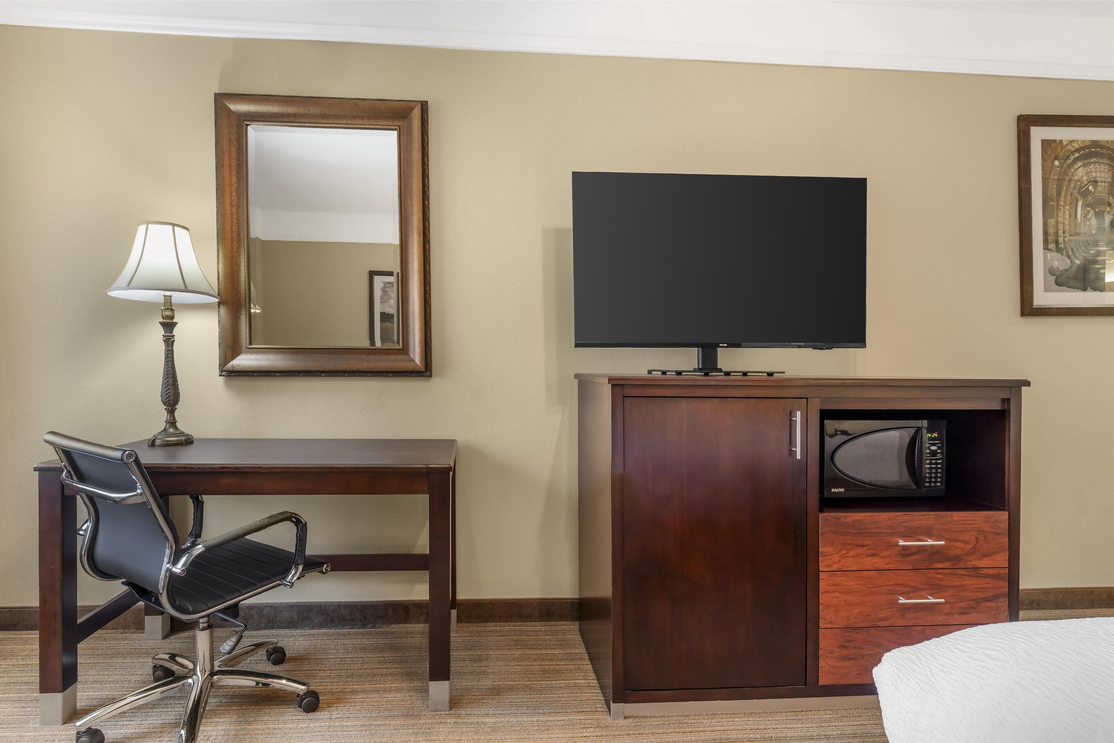 A hotel room with a wooden desk, a chair, a television, a microwave, and a bed.