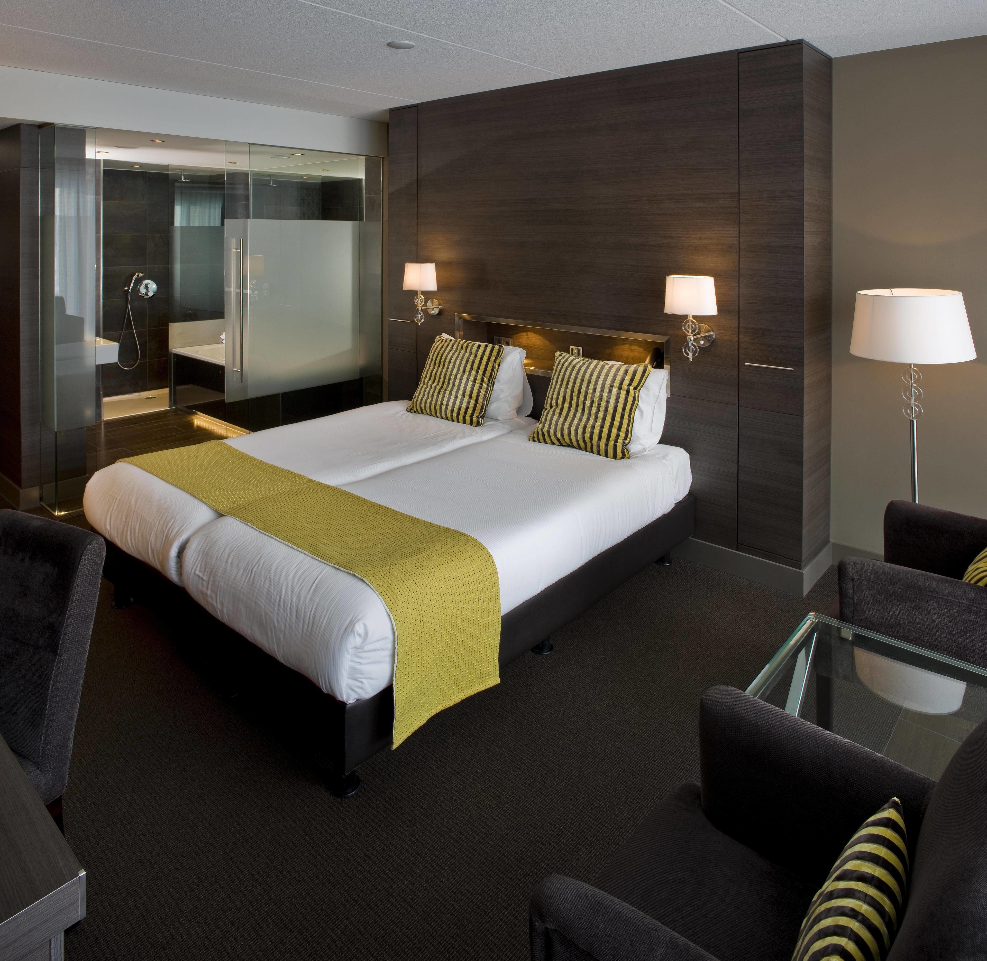 A modern hotel room with a large bed, two bedside lamps, a glass shower enclosure, and a seating area with a glass table and chairs.