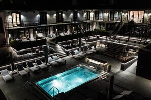 Un área de piscina en la azotea con sillones de descanso y una piscina, rodeada por un edificio de varios niveles con asientos exteriores y escaleras.