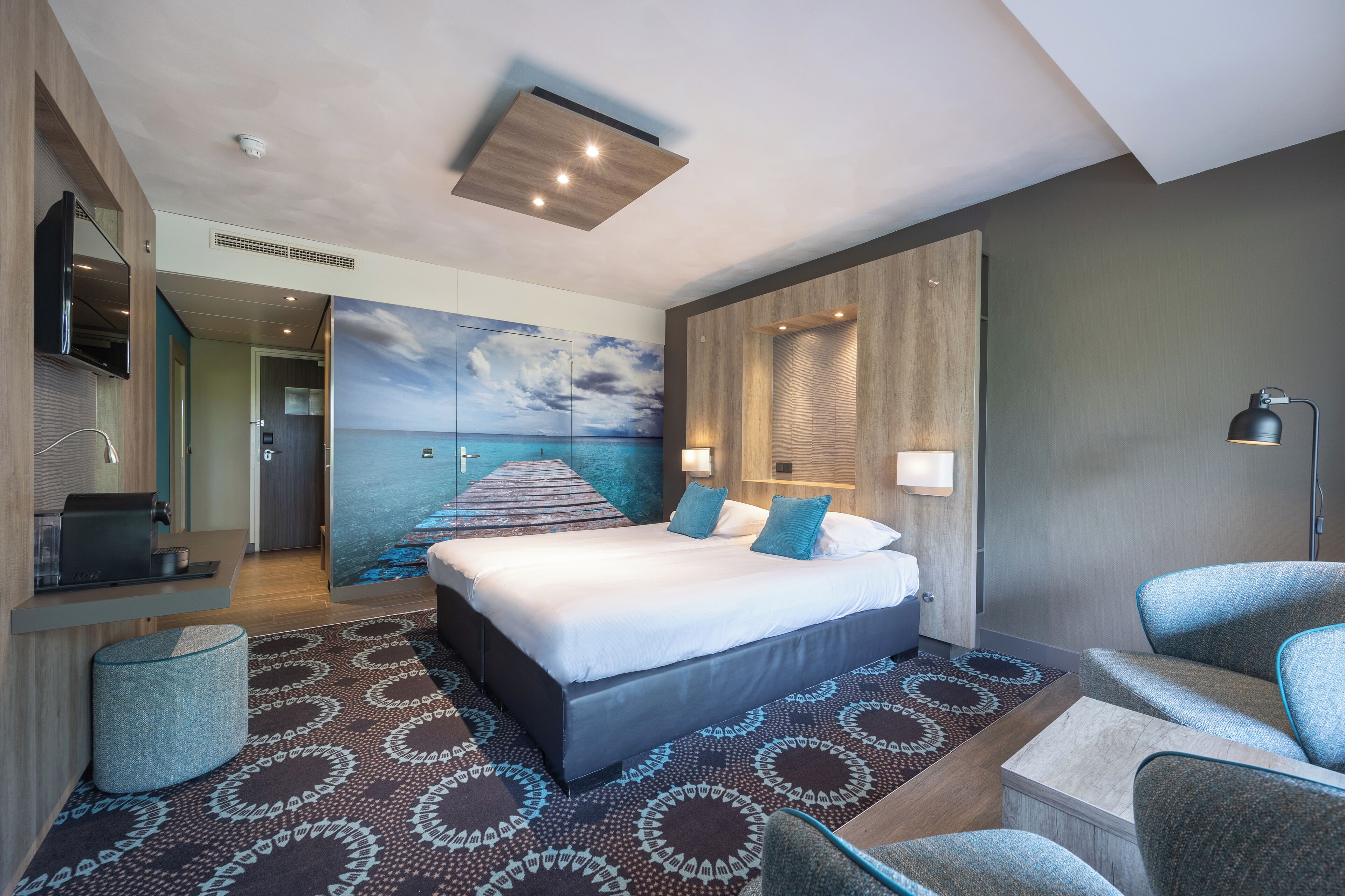 Una habitació d'hotel moderna amb un llit gran, un escriptori, un televisor i un mural d'una escena de platja.