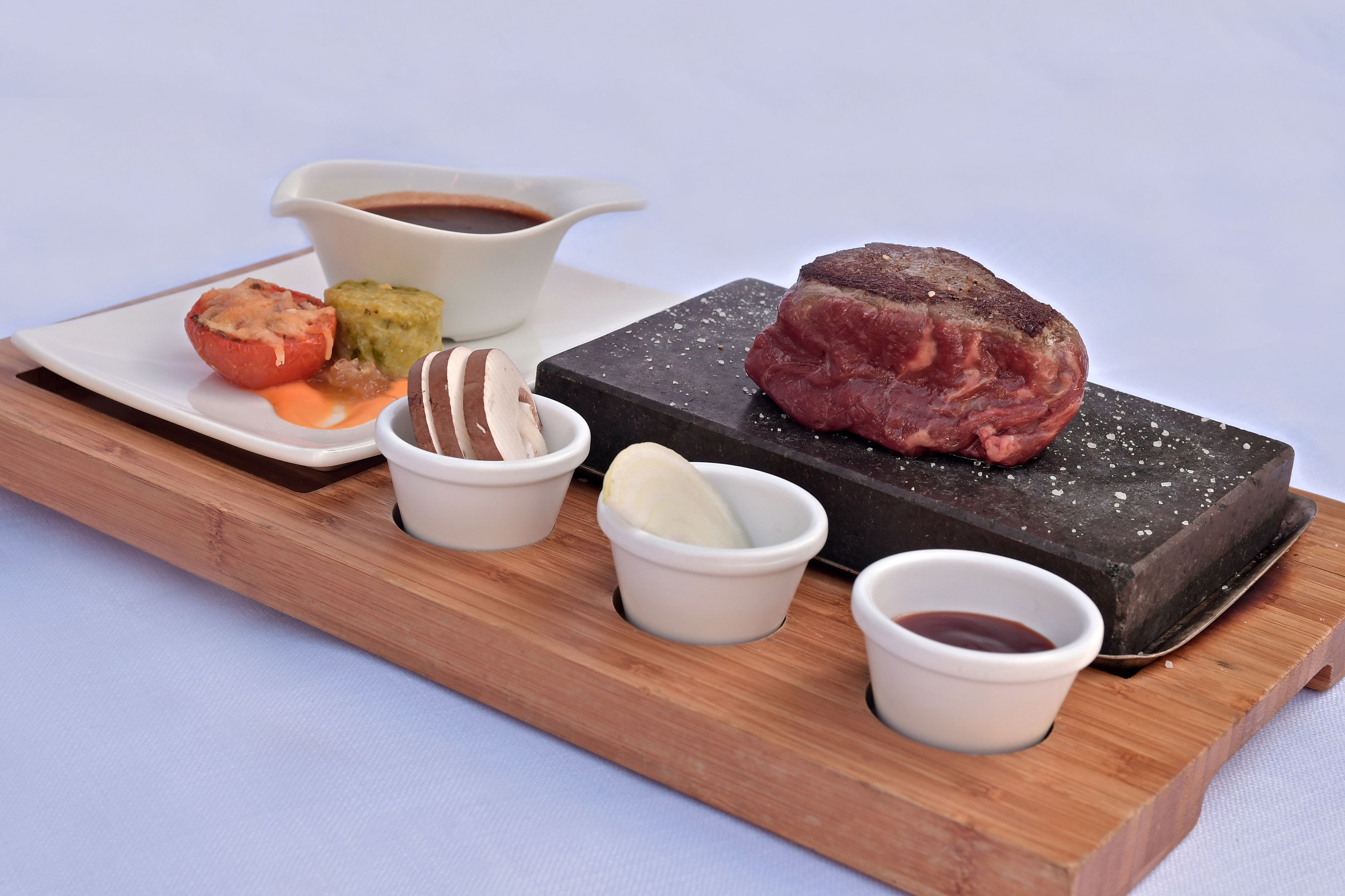 Une planche de service en bois sur laquelle on a disposé un steak, une tomate rôtie, un légume vert et de petits ramequins de sauces.