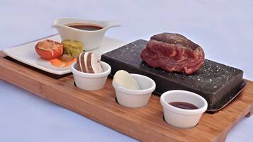 A wooden serving board with a steak, a baked tomato, a green vegetable, and small bowls of sauces.