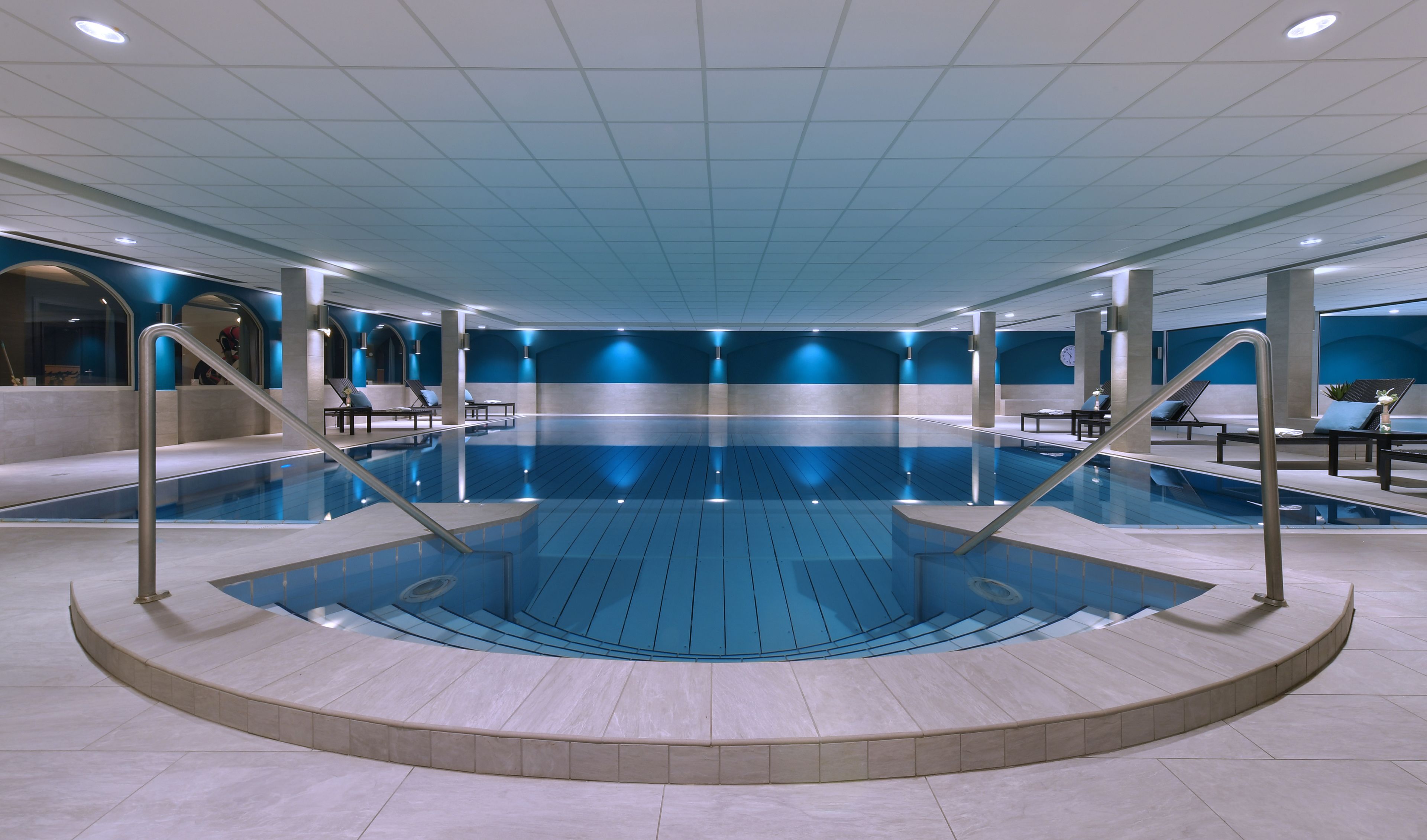 Una amplia piscina interior con una zona de trampolín en el centro, rodeada de sillones de descanso y suelo de baldosas.