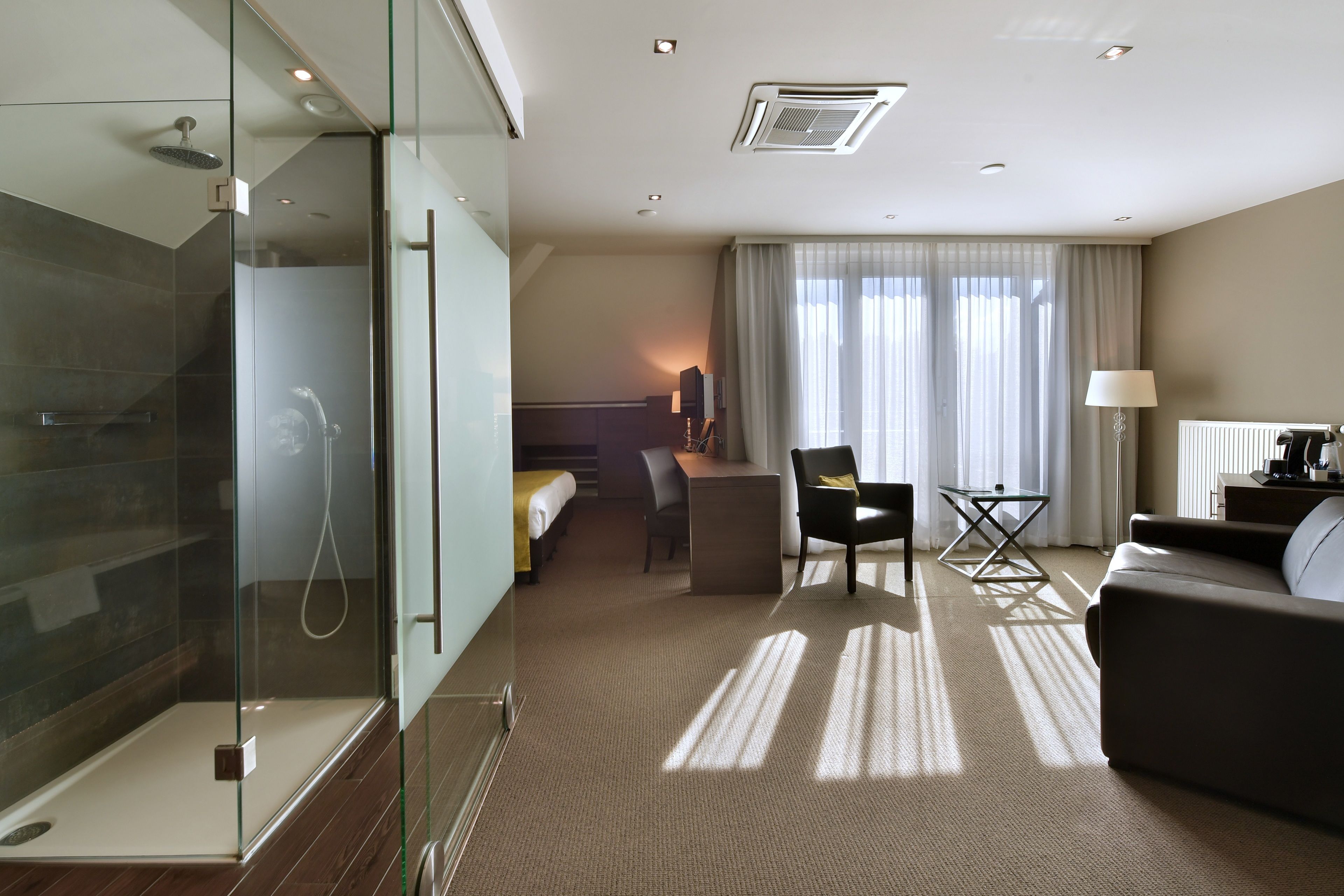 Een moderne hotelkamer met een douchecabine van glas, een bed, een bureau, een stoel en een bank.
