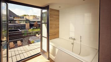 Een moderne badkamer met een groot bad, een glazen deur naar een balkon en uitzicht op de gebouwen.