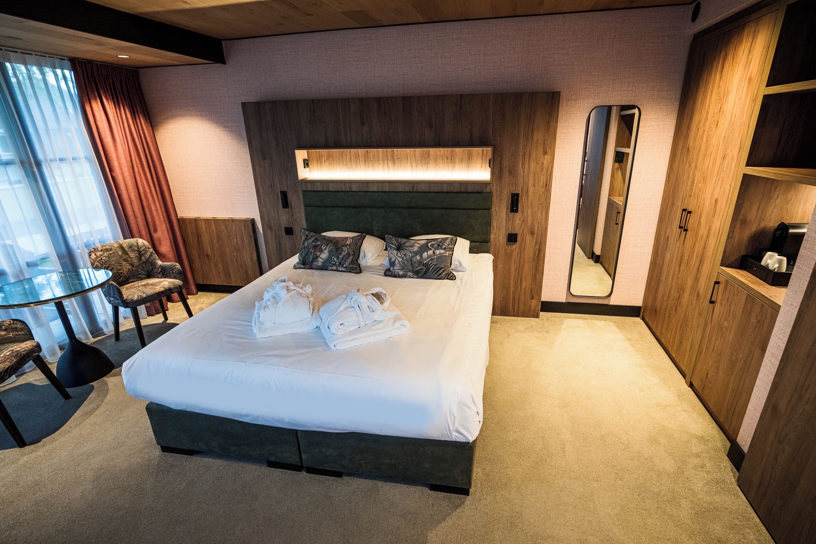 A hotel room with a large bed, a round table with chairs, a mirror, and wooden cabinets.
