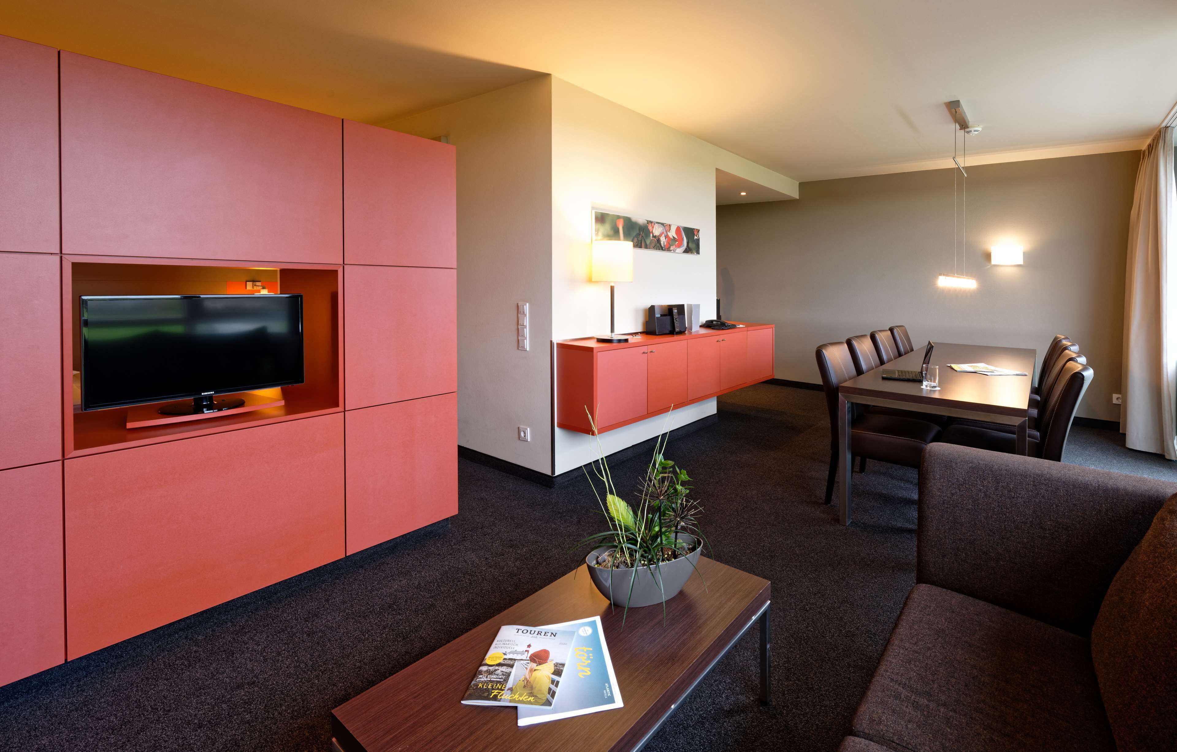A modern hotel room with a flat-screen TV, a dining table with chairs, a sofa, a coffee table with a magazine, and a potted plant.