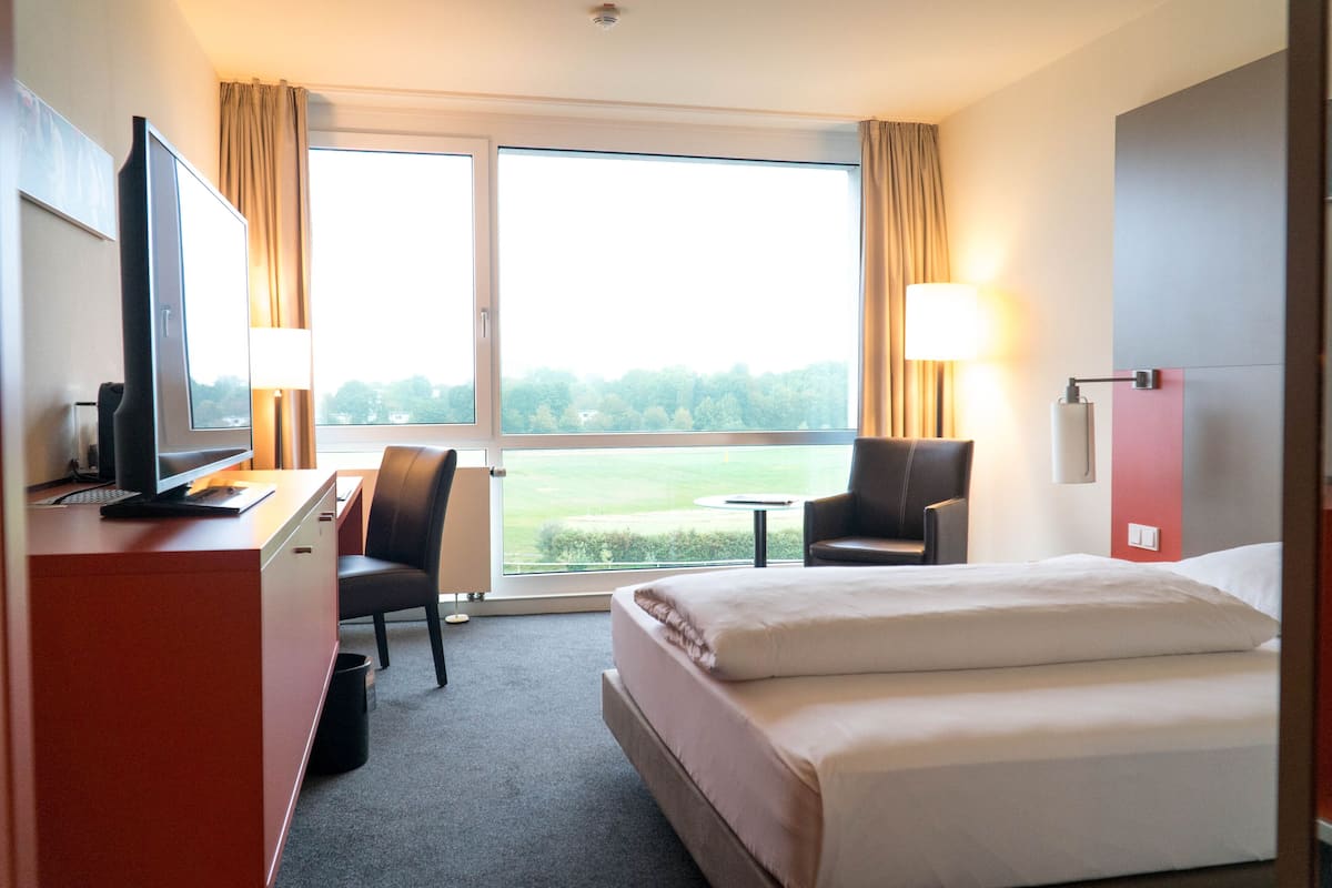 Habitación de hotel con cama, escritorio, silla, televisor y ventanas amplias.