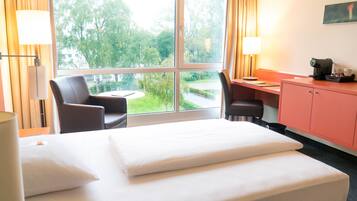 A hotel room with a bed, desk, chair, and a view of greenery outside.