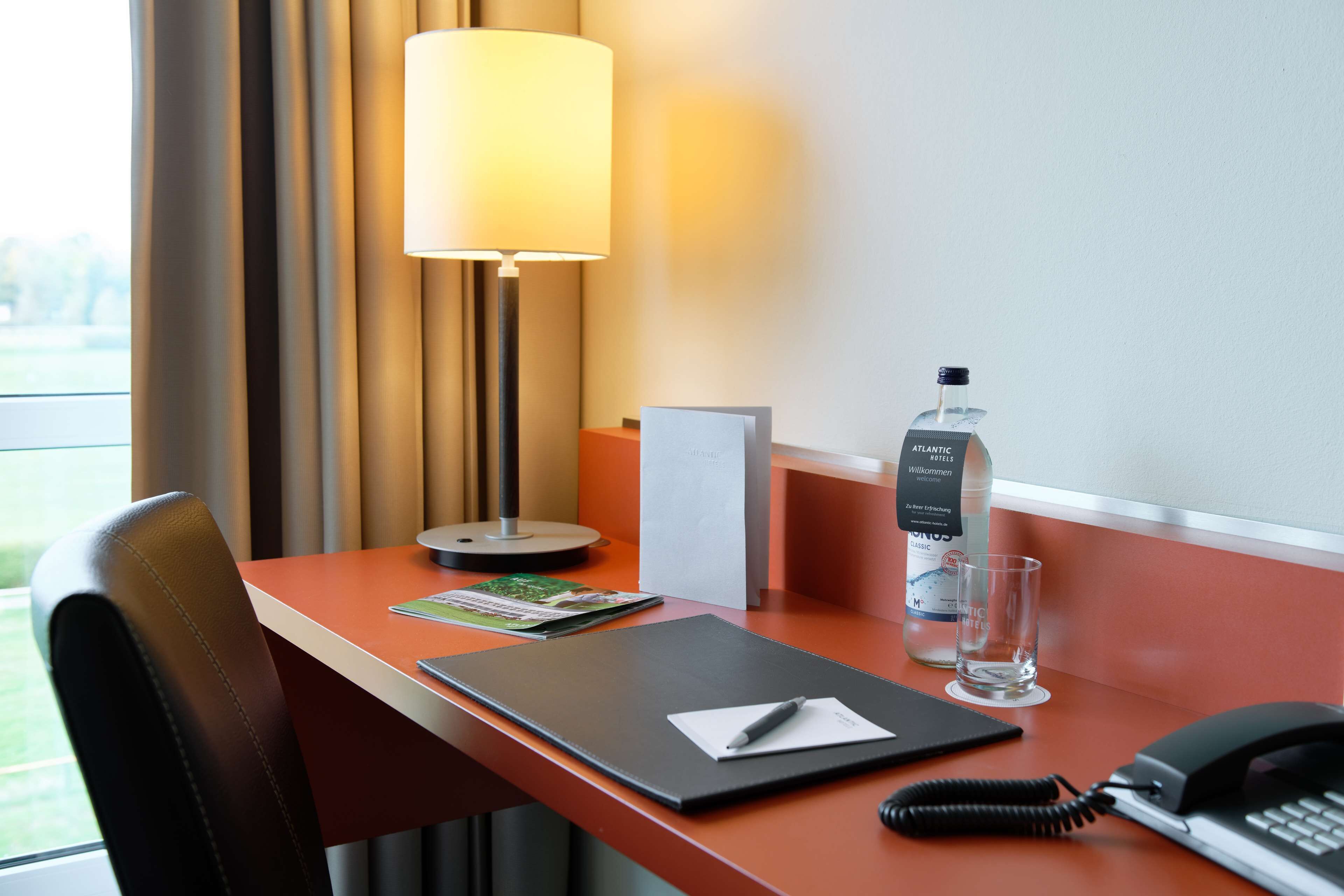 A hotel room desk with a lamp, a bottle of water, a glass, a phone, and a notebook.