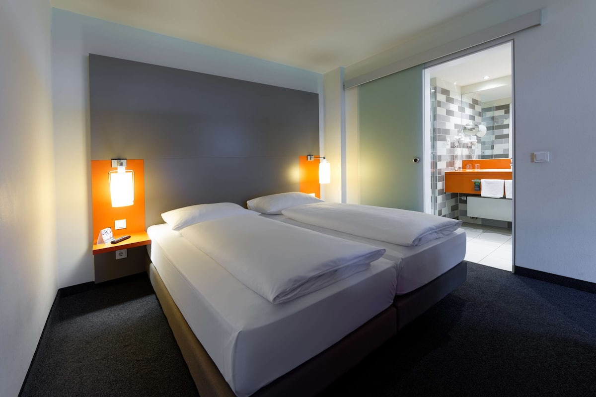 Habitación de hotel con dos camas, una mesita de noche y un baño visible a través de una puerta abierta.