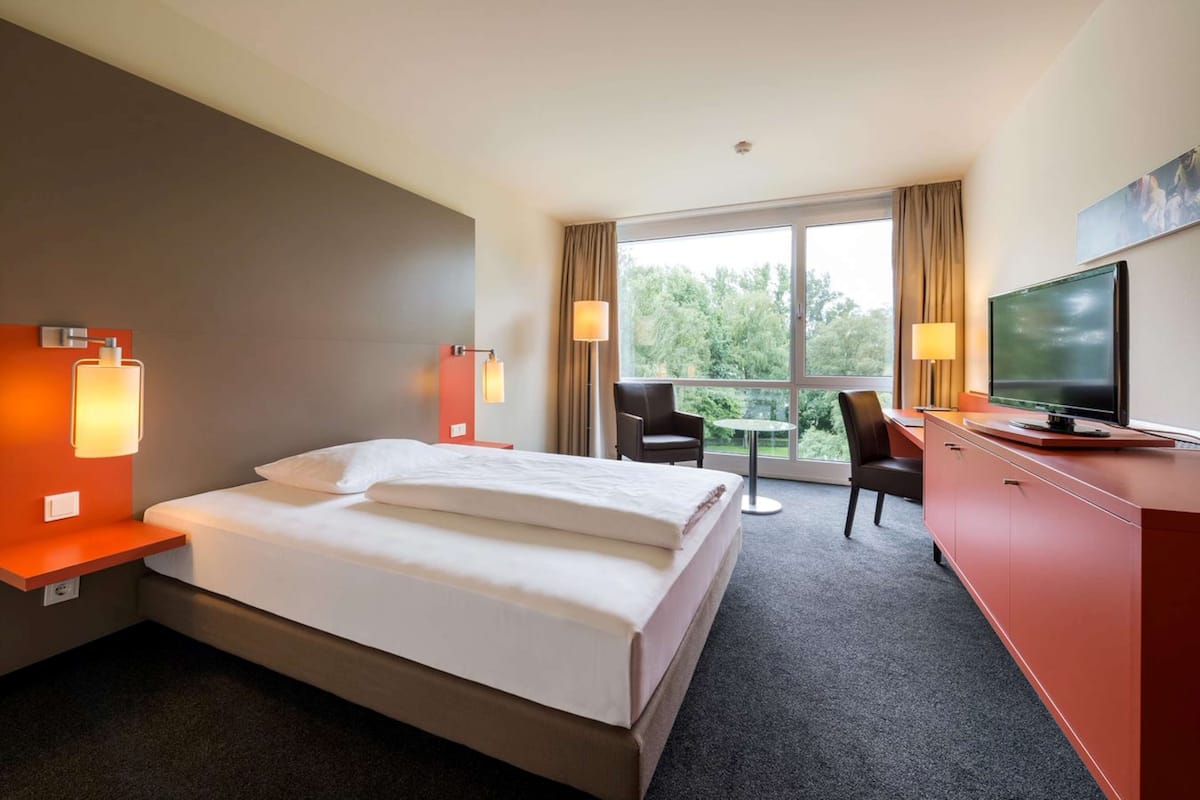 Habitación de hotel con cama, escritorio con televisor, silla y vista a los árboles por la ventana.