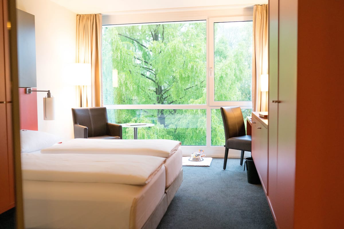 Habitación de hotel con una cama grande, dos sillas, una mesita y una ventana con vista a árboles.