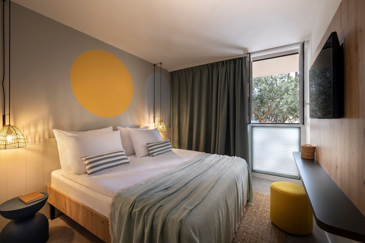 A modern hotel room with a large bed, a round yellow wall decoration, a window with curtains, and a flat-screen TV mounted on the wall.