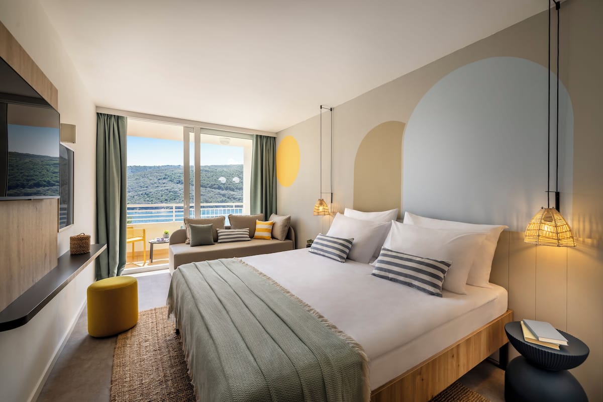 A modern hotel room with a large bed, a sofa, a TV, and a view of the landscape.