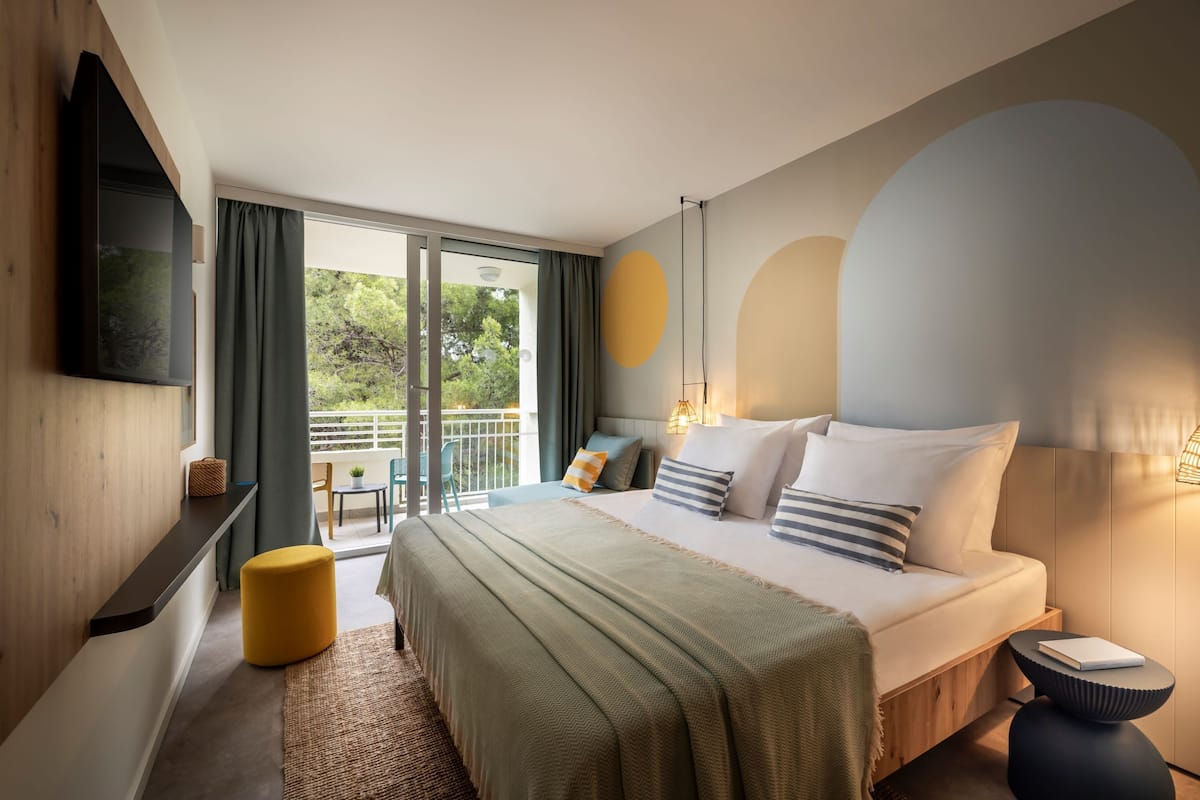 A modern hotel room with a large bed, a flat-screen TV, a balcony with a view of greenery, and a minimalist decor style.