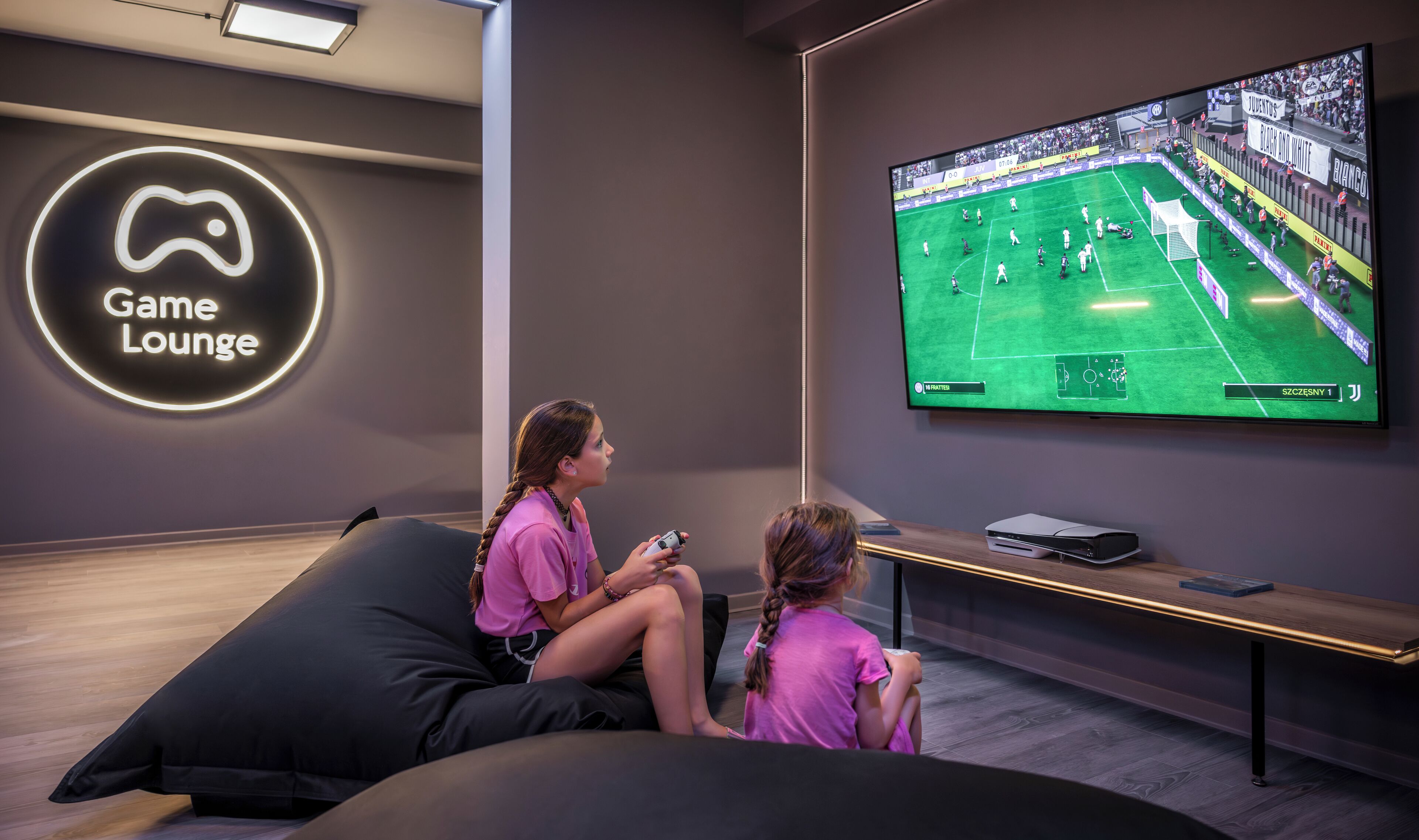 Un espace de détente moderne doté d’un grand écran diffusant un match de football, de sièges confortables et d’un panneau néon indiquant un espace dédié aux jeux.