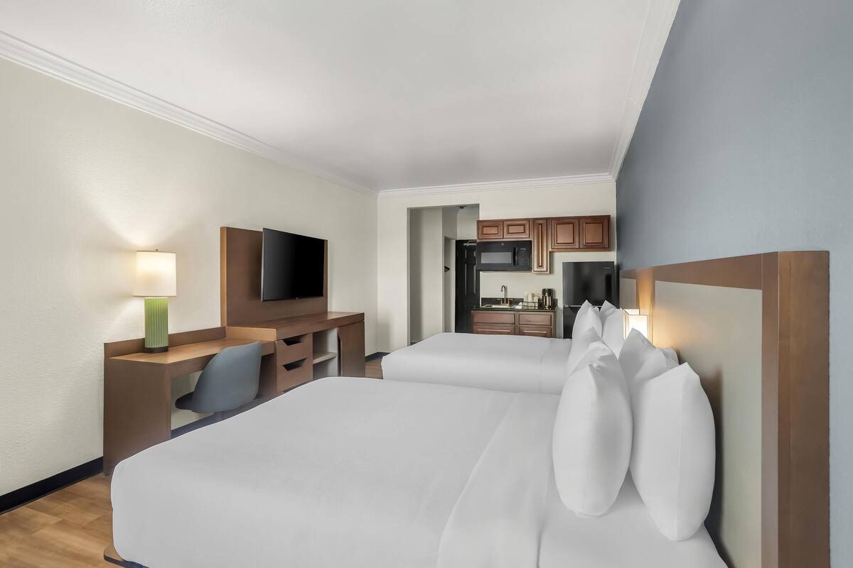 Quarto de hotel com duas camas, uma escrivaninha com cadeira, televisão e cozinha compacta.