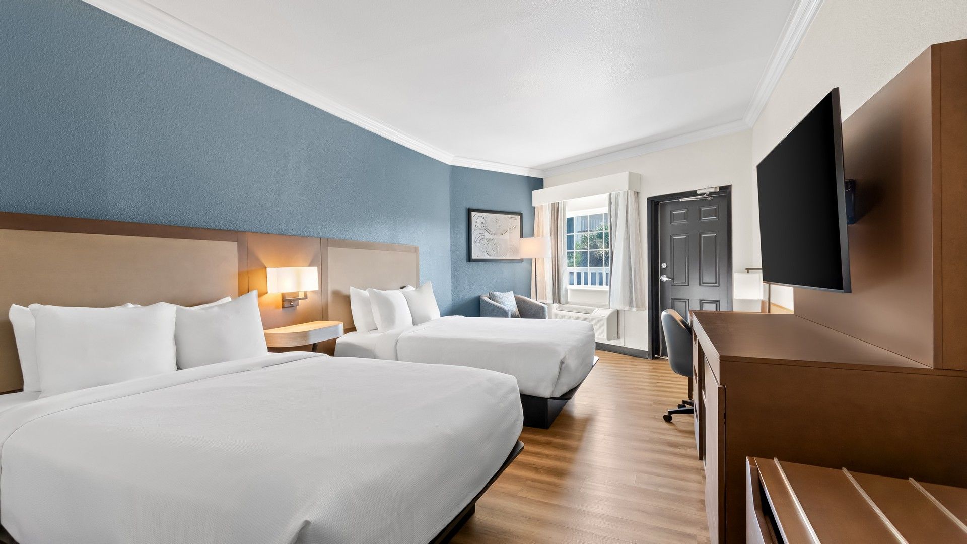 A hotel room with two beds, a desk, a chair, a TV, and a window.